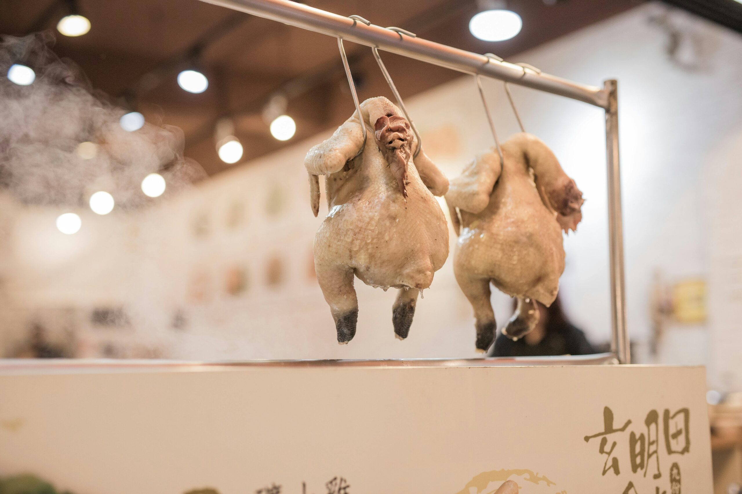 Two hanging chickens being prepared at a market stall indoors with steam visible.